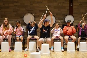 A group of kids play bucket drums under the direction of a camp leader. They hold their sticks up, ready to play.